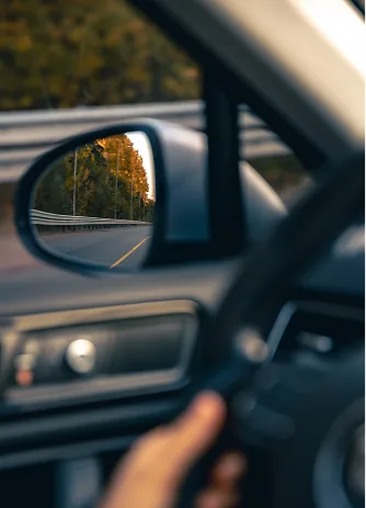 A driver’s hand on the steering wheel, with an autumn road reflected in the wing mirror